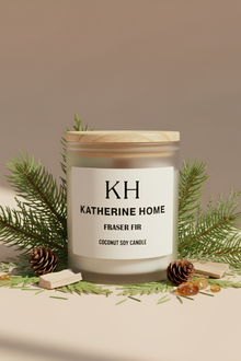 Candle with a wooden lid and 'KH Katherine Home' label, surrounded by greenery on a beige background
